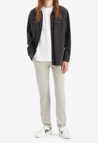 Ciemnoszara koszula z guzikami nałożona na białą koszulkę, w zestawie z jasnobeżowymi spodniami i białymi trampkami z granatowymi akcentami. Swobodny design stroju.