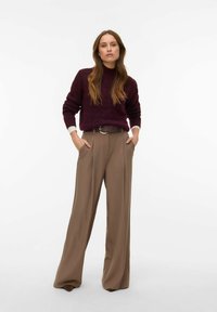 Bordowy sweter z dzianiny z wysokim kołnierzem. Beżowe spodnie o szerokich nogawkach z paskiem z srebrną sprzączką. Modelka pozuje z rękami w kieszeniach.