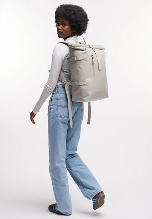 Sac à dos gris à rabat avec des bretelles ajustables, une poche zippée à l'avant et des boucles sur les côtés. Porté sur le dos par une personne vêtue de vêtements légers et en denim.