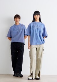 Två individer står sida vid sida. Båda bär överdimensionerade, blå bomullströjor med broderade mönster. Den ena har svarta byxor, den andra ljusbeiga cargobyxor.