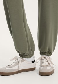 Olivově zelené tepláky s elastickými manžetami v kombinaci se světle šedými a bílými teniskami, které mají černou patu a béžové semišové detaily.
