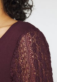 Bordový top s jednobarevným tělem a krajkovými rukávy s propracovanými květinovými vzory a vlnitými okraji.