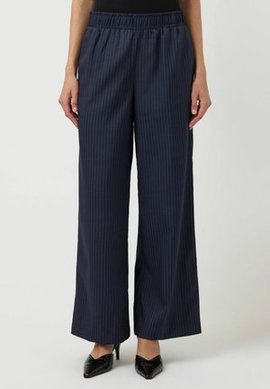 Kvinne kledd i marineblå vidbukser med smale striper og elastisk midje, samt svarte spisse hæler, stående foran en ensfarget bakgrunn.