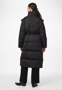Lang svart puffjakke med belte i midjen, med høy krage og horisontale quiltede seksjoner. Materialet ser tykt og isolert ut.