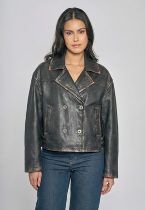 Femme aux longs cheveux noirs portant une veste vintage en cuir noir à double boutonnage et un jean bleu, debout devant un fond gris uni.