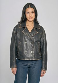 Kvinde med langt mørkt hår iført en vintage sort læder-dobbeltbrystet jakke og blå jeans, stående mod en ensfarvet grå baggrund.