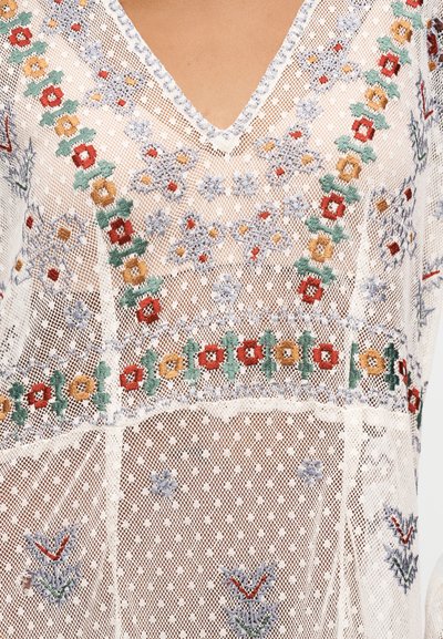 Haut en maille blanche avec broderie florale colorée en rouge, vert, jaune et bleu, présentant un col en V et des détails de motifs complexes.