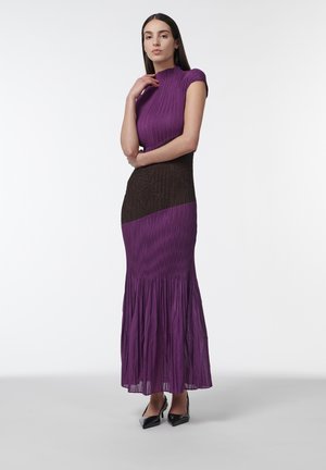 Mujer de pie, vestida con un vestido corto de manga corta en tonos púrpura y negro con textura, cuello alto y bajo plisado, combinado con tacones negros de punta afilada.