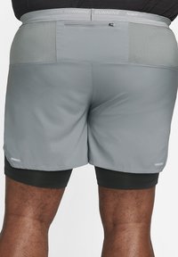 Sive Nike tekaške kratke hlače z lahkim materialom, mrežastimi detajli na pasu in odsevnimi podrobnostmi na robu. Črne spodnje hlače so vidne.