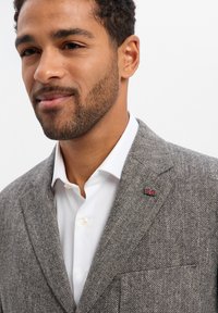 Szary blezer w jodełkę z klapami z wycięciem, zestawiony z białą koszulą. Na klapie znajduje się mała przypinka z flagą Wielkiej Brytanii. Miękka tekstura.