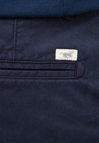Navy cotton pants feature a double pleat pocket with a stitched label displaying a horse design in gray on a white background.