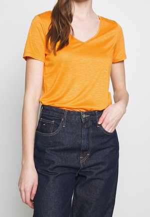 Woman wearing bright orange short-sleeve V-neck T-shirt tucked into high-waisted dark blue jeans, with one hand in pocket.