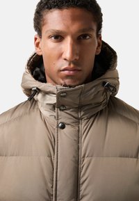 Beige pufferjack met een capuchon, met een quilted ontwerp, drukknoopsluiting en trekkoordaccenten bij de kraag. Soepele textuur.