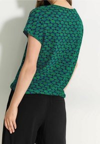 Kortærmet grøn bluse med marineblå blomsterdesign, rund udskæring og en afslappet pasform, parret med sorte bukser. Blødt stof.