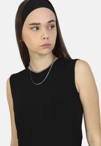 Top negro sin mangas con una textura suave, combinado con dos collares de cadena plateados. La modelo lleva una diadema negra y tiene el cabello lacio castaño.