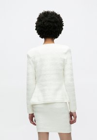 Λευκή υφασμάτινη δύο τεμαχίων ένδυση που περιλαμβάνει στενή μίνι φούστα και ραμμένο σακάκι με πεπλούμ και μακριά μανίκια.