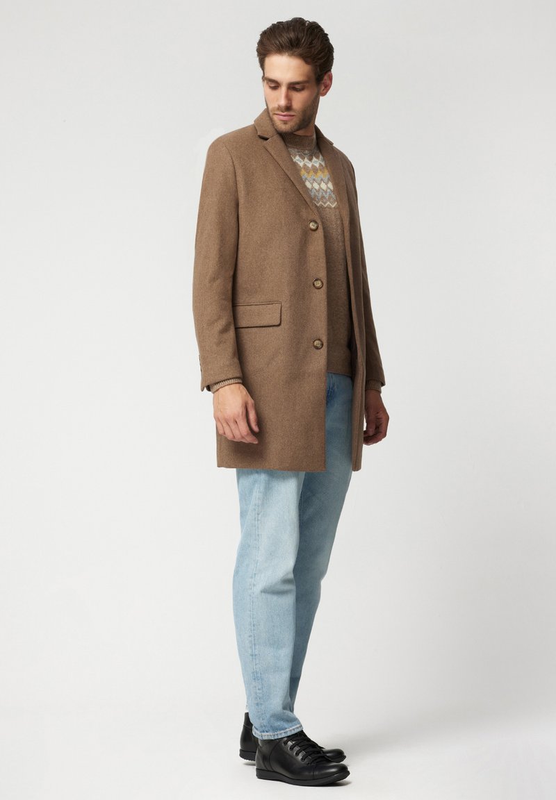 Brązowy płaszcz z mieszanki wełny z klapami, dwoma przednimi kieszeniami i guzikami, w zestawie z jasnoniebieskimi dżinsami i czarnymi butami. Włożony sweter w wzory.