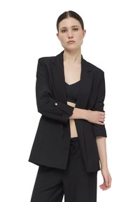 Czarna marynarka dopasowana, z jednopiersiowym krojem, wyraźnymi ramionami i podciągniętymi rękawami; noszona na czarnej bluzce.