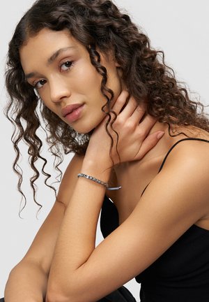 Silbernes Armband mit Buchstaben auf einem blauen Band, getragen am Handgelenk. Das Modell hat lockiges Haar, trägt ein schwarzes ärmelloses Oberteil und steht vor einem neutralen Hintergrund.