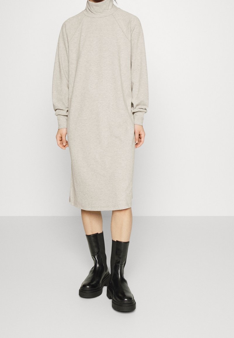 Personne portant une robe midi à manches longues col roulé beige clair avec des fentes et des bottes noires épaisses mi-mollet, debout sur un sol gris.