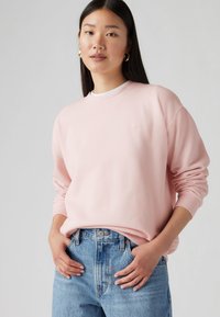 Sweatshirt rose clair avec un col et des poignets côtelés, arborant un petit logo sur la poitrine, associé à un jean en denim bleu taille haute.