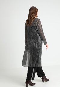 Lang, gennemsigtig jakke med lodrette metalliske striber i sort og sølv, med lange ærmer, åben foran og afslappet pasform.