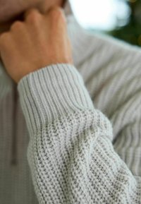 Personne en pull gris clair tricoté, reposant le menton sur la main, gros plan montrant les détails du tissu texturé de la manche et du poignet.