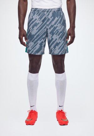 Un modèle se tient debout, vêtu d’un short de football gris à motifs, d’un maillot blanc et de chaussettes montantes jusqu’aux genoux, avec des chaussures de football orange vif.