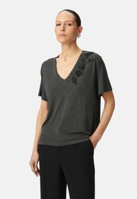 T-shirt col V gris foncé en tissu doux à manches courtes. Présente un ornement floral en sequins noirs sur une épaule.
