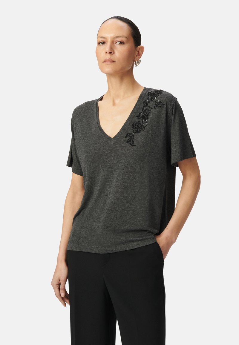 T-shirt col V gris foncé en tissu doux à manches courtes. Présente un ornement floral en sequins noirs sur une épaule.