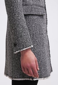 Gros plan sur la main d’une personne reposant à son côté, portant un manteau en tweed texturé noir et blanc avec des bords effilochés.