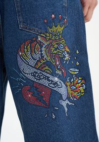 Denim jeans featuring a colorful rhinestone design of a tiger and crown, with textured embellishments in red, yellow, and blue.