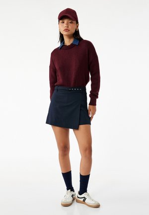 Suéter burdeos, camisa de cuello azul marino, falda envolvente azul oscuro, calcetines azules marinos, zapatillas blancas con detalles negros, completan el atuendo.