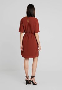 Robe couleur rouille avec des manches courtes, taille ceinturée, détail en forme de trou de clé au dos, et une longueur jusqu'aux genoux. Portée avec des escarpins noirs à brides.