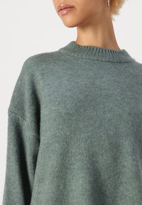 Maglione verde in lana con scollo a carreggiata a costine e spalle basse. Texture morbida, design semplice, vestibilità casual, che mostra la parte superiore del capo.