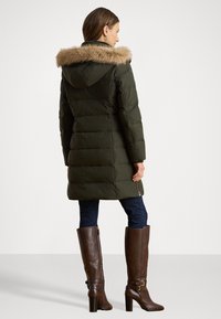 Giacca puffa verde oliva con cappuccio foderato in pelliccia sintetica, caratterizzata da un design trapuntato e due cerniere laterali, abbinata a stivali marroni fino al ginocchio.