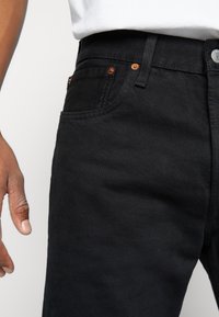 Jeans de mezclilla negra con un diseño ajustado, que presentan remaches de cobre, costuras destacadas y una textura suave. Mano colocada al lado.