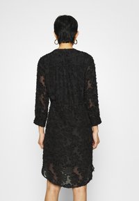 Vestido negro con tela transparente, mangas tres cuartos y bordado floral texturizado. El dobladillo cae ligeramente por encima de la rodilla.