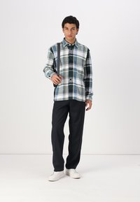 Plaid button-up shirt in green, white, and navy; relaxed fit; paired with dark pants and white sneakers; model holds a shoulder bag.