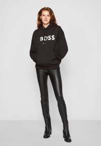 Zwarte hoodie met een wit "BOSS" logo, voorzak, en een capuchon met trekkoord; gecombineerd met strakke zwarte neplederen leggings en zwarte enkellaarsjes.