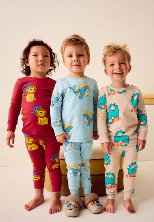 Tre bambini in piedi al chiuso che indossano pigiami colorati a tema animali: leoni rossi, balene blu con pantofole e dinosauri beige, sorridenti e in posa.