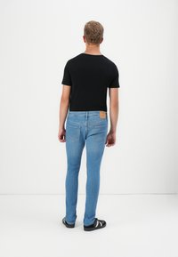 Camiseta negra de manga corta para hombres combinada con jeans ajustados de color azul claro. Los jeans presentan un parche marrón en la cintura trasera y en los bolsillos traseros.