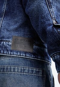 Džinsinis švarkas, pasiūtas iš tekstūrinio, tamsiai mėlyno audinio. Gale yra juodos odos emblema su iškilmingu tekstu. Matomi siuvimo detalės.