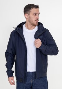 Granatowa kurtka z kapturem o gładkiej powierzchni, z zamkiem błyskawicznym i regulowanymi sznurkami. Posiada logo na klatce piersiowej oraz boczną kieszeń.