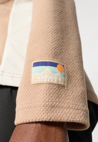 Beige fleece takki hihna, jossa on teksturoitu pinta. Värikäs päällyste esittelee vuoria ja auringon, ja siinä on teksti "IC3PAK."