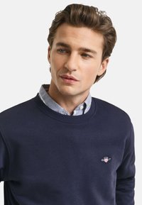 Marine crewneck sweatshirt met ribbelmanchetten, gedragen over een lichtblauw overhemd met kraag. Heeft een klein logo op de borst.