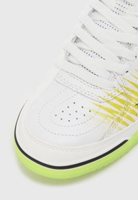 Sneaker blanc avec une tige en cuir texturé, accents dégradés jaunes, boîte à orteils perforée et semelle translucide verte avec bord noir.