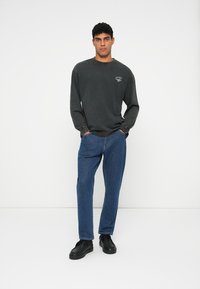 Mørkegrå langermet skjorte med et lite grafisk motiv på brystet, kombinert med blå jeans og svarte joggesko. Enkelt, uformelt design.