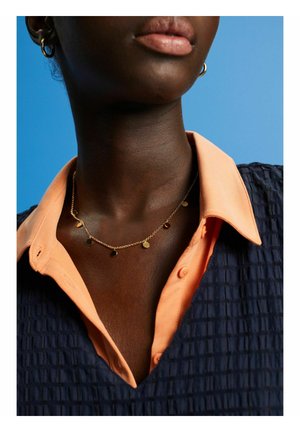 Gouden ketting met ronde bedels, gedragen over een perzikkleurig overhemd met kraag en een textuurvol donkerblauw topje, die de gelaagde ontwerpelementen benadrukt.