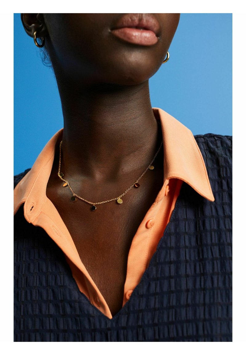 Collana dorata con pendenti circolari, indossata sopra una camicia color pesca con colletto e un top blu navy testurizzato, che mette in evidenza i dettagli del design a strati.